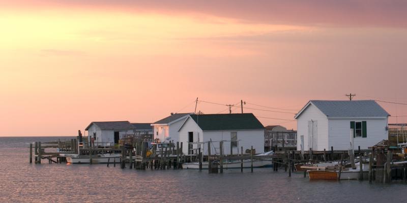 Beach houses on stilts overhanging the water with pink hues of dawn in the background in Tangier Island, Virginia, USA
