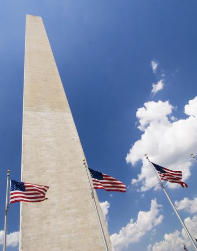 A picture of the Washington Monument from the ground looking up towards the blue, cloud-dotted sky. There are American flags flying around the base of the monument